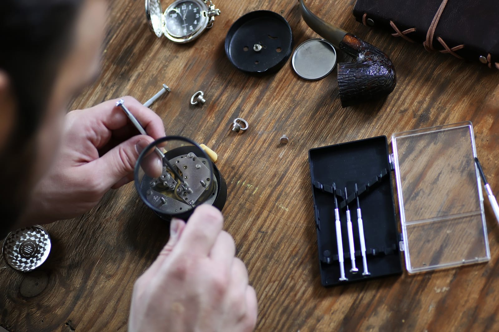 Orologiaio che osserva con una lente un movimento a orologeria aperto su un banco di legno, tra strumenti e una borsa di cuoio
